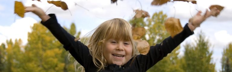 Ein kleines Mädchen wirft Herbstblätter in die Luft und lächelt freudig.